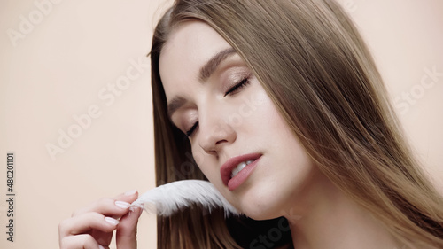 sensual young woman with closed eyes holding feather near face isolated on beige.