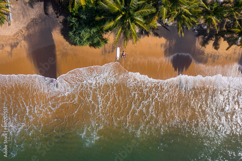 Aerial photo of surfer lying next to surfboard on sandy ocean shore in STAR pose and sun tanning under tall palm trees. Soft waves washing his legs on Sri Lank beach island. Exotic vacation concept photo