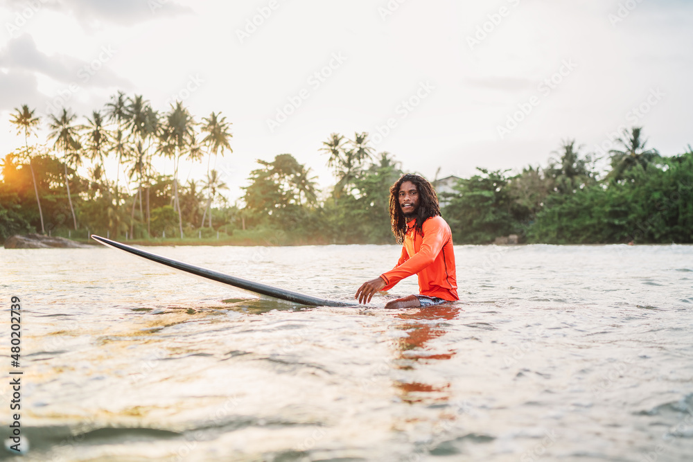 Black long-haired teen man floating on long surfboard, waiting for a wave ready for surfing with palm grove litted sunset rays. Extreme water sports and traveling to exotic countries concept. Black long-haired teen man floating on long surfboard, waiting for a wave ready for surfing with palm grove litted sunset rays. Extreme water sports and traveling to exotic countries concept.