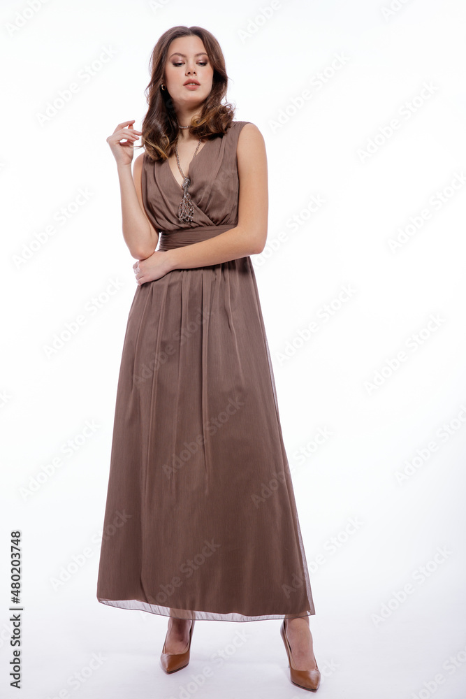 High fashion photo of beautiful elegant young woman in a pretty beige brown dress with a neckline, necklace posing on white background. Slim figure, make up,  luxurious hair, shiny curls. Studio Shot