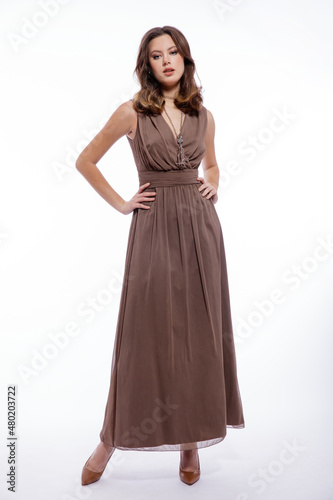 High fashion photo of beautiful elegant young woman in a pretty beige brown dress with a neckline, necklace posing on white background. Slim figure, make up,  luxurious hair, shiny curls. Studio Shot