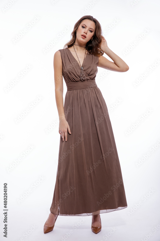 High fashion photo of beautiful elegant young woman in a pretty beige brown dress with a neckline, necklace posing on white background. Slim figure, make up,  luxurious hair, shiny curls. Studio Shot