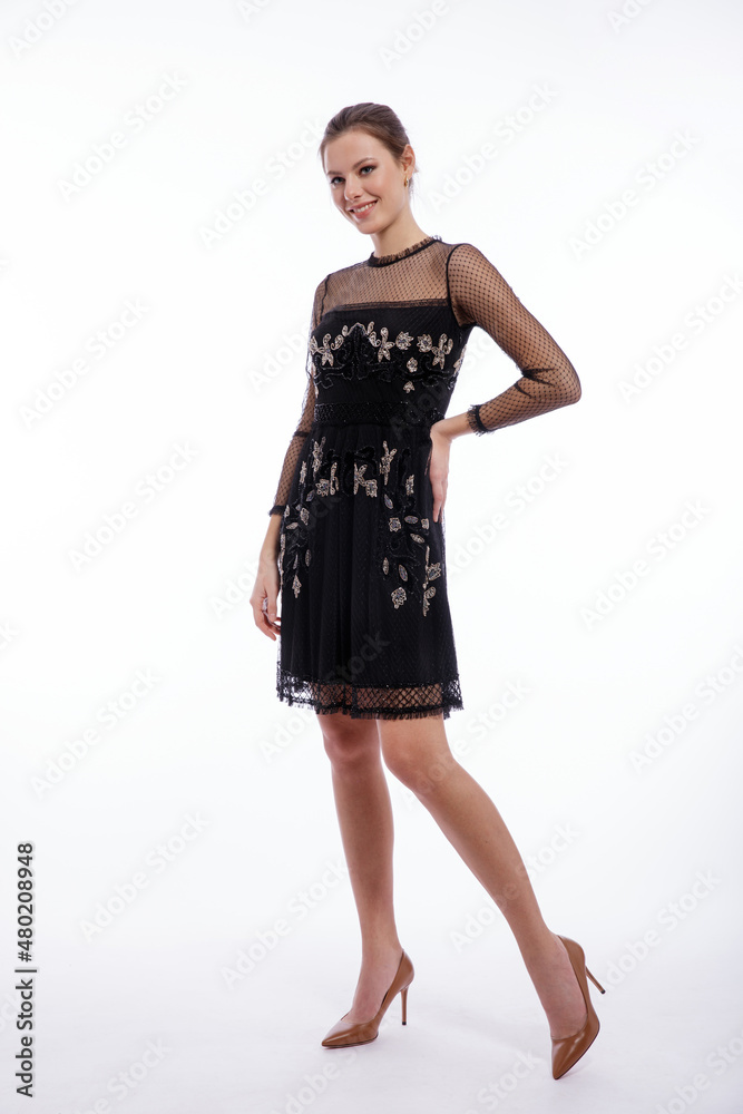 Fototapeta premium High fashion photo of a beautiful elegant young woman in a pretty black dress with lace posing on white background. Studio shot. Slim figure, hair is gathered in a ponytail