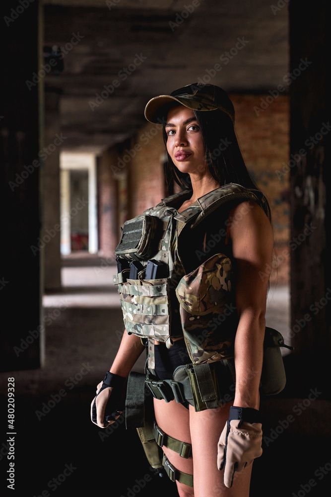 Powerful woman in military gear posing indoors in abandoned building. brunette fit female in shorts is looking at camera confidently. warfare, military forces, defense concept. close-up portrait