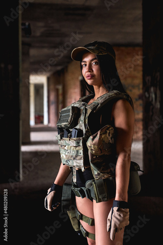 Powerful woman in military gear posing indoors in abandoned building. brunette fit female in shorts is looking at camera confidently. warfare  military forces  defense concept. close-up portrait