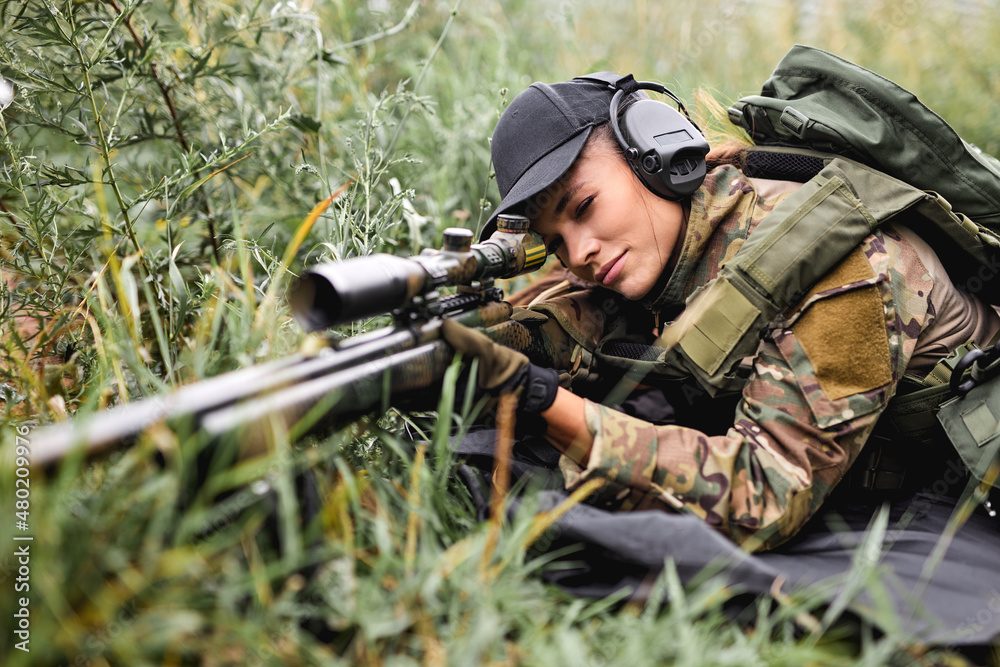beautiful focused woman with gun weapon with suppressor in uniform lying on ground on grass in field in shelter, expect the enemy. caucasian military lady in headset, gear aiming rifle, ready to shoot