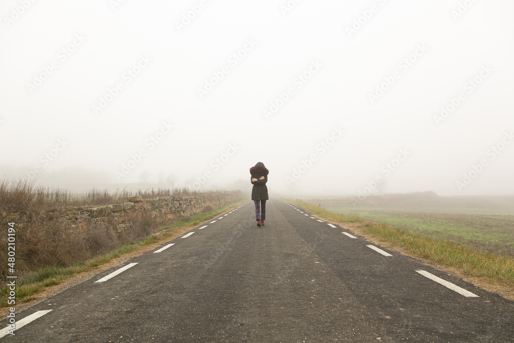 Chica con frío con abrigo caminando con niebla por una carretera secundaria,  huida 