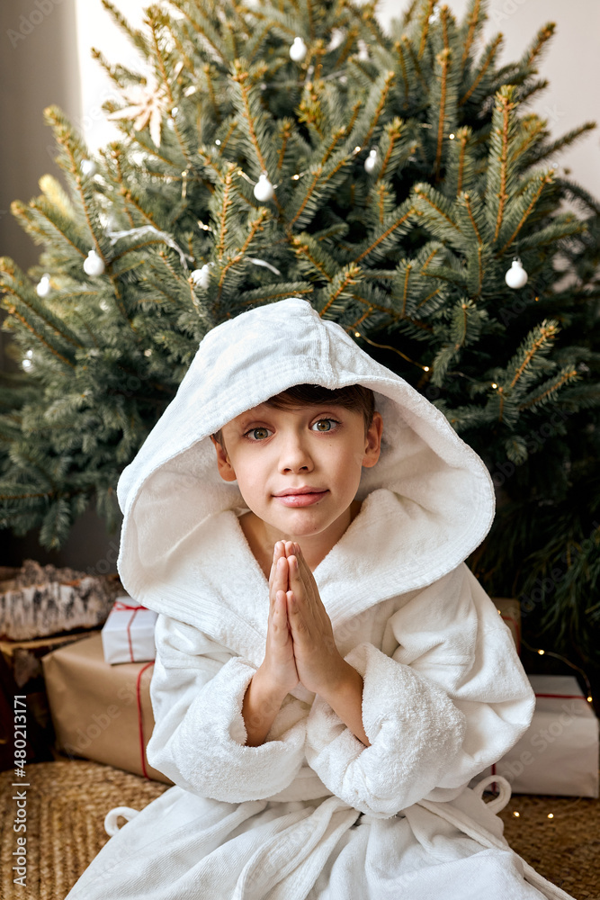 Obraz premium American Child Making Magic Wish for Winter Holidays. Boy Near the Christmas Tree at Home. Teen Boy In Bathrobe Praying. Waiting for Santa at Night. Holidays, Weekends, New Year, Christmas 2022