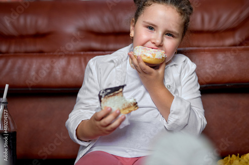 hungry caucasian girl is eating Unhealthy sweet cakes and watching tv sitting on floor next to sofa. hungry child is eating tasty donut and cake  biting donut at home in a hurry. Unhealthy fast food.