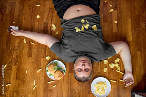 Fényképezés corpulent fat boy eat junk food while lying on floor at home, with french fries and chips
