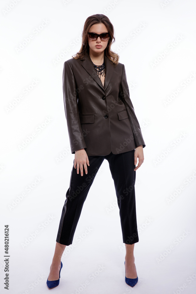 High fashion photo of beautiful elegant young woman in a pretty suit, leather dark olive khaki jacket, trousers, pants, stylish sunglasses posing on white background. Studio shot. Slim figure High fashion photo of beautiful elegant young woman in a pretty suit, leather dark olive khaki jacket, trousers, pants, stylish sunglasses posing on white background. Studio shot. Slim figure