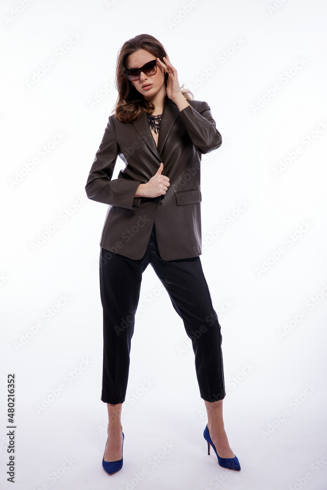 High fashion photo of beautiful elegant young woman in a pretty suit, leather dark olive khaki jacket, trousers, pants, stylish sunglasses posing on white background. Studio shot. Slim figure High fashion photo of beautiful elegant young woman in a pretty suit, leather dark olive khaki jacket, trousers, pants, stylish sunglasses posing on white background. Studio shot. Slim figure