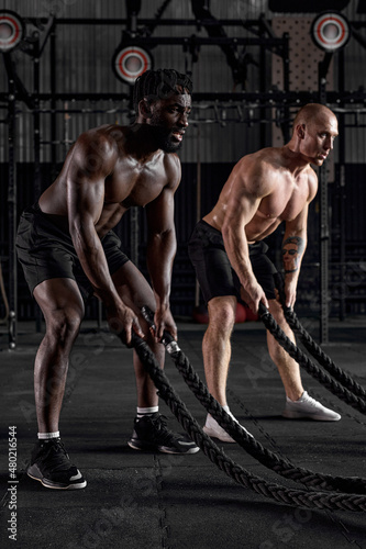 Side View On Young Diverse Shirtless Men With Battle Rope Doing Exercise In Functional Training Fitness Gym. Caucasian And Afro American Guys During Intense Training. In Dark Sports Club