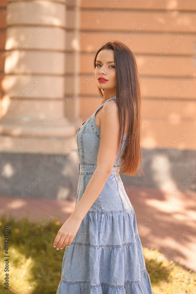 Cheerful long haired brunette lady model in casual denim sundress with red lipstick poses for camera on city street on summer day side view