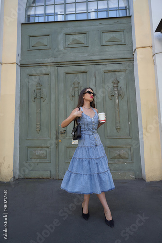 Stylish brunette woman wearing long denim summer dress holds cup of drink standing near building with vintage door low angle shot