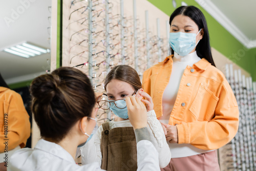 blurred ophthalmologist trying eyeglasses on girl in medical mask near asian woman in optics store.