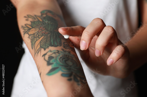 Fototapeta Woman applying cream on her arm with tattoos against black background, closeup