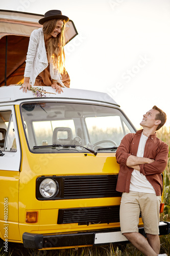 Positive caucasian couple spend time in nature, came there on mini van, woman is standing on roof of yellow van while man is standing next to van. hippie life, travel, holidays at sumemr photo