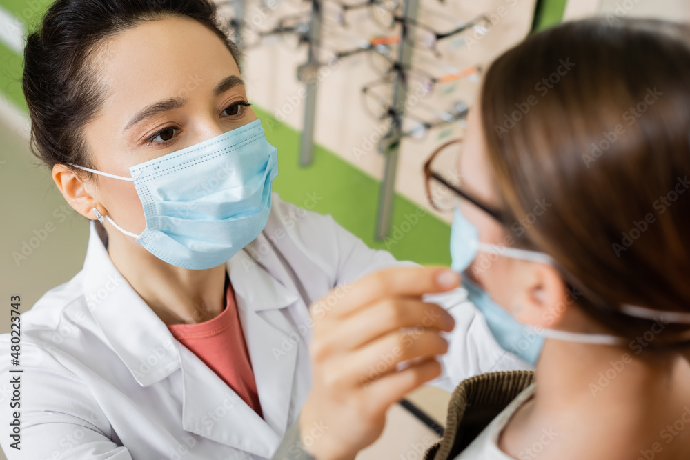 oculist in medical mask trying eyeglasses on blurred girl in optics store. oculist in medical mask trying eyeglasses on blurred girl in optics store.