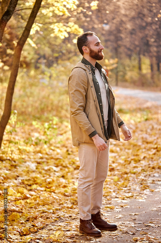 Trendy handsome man posing in autumn park alone, looking at side smiling, wearing stylish clothes. Caucasian bearded guy in coat enjoy the walk during leaf fall in forest. People concept
