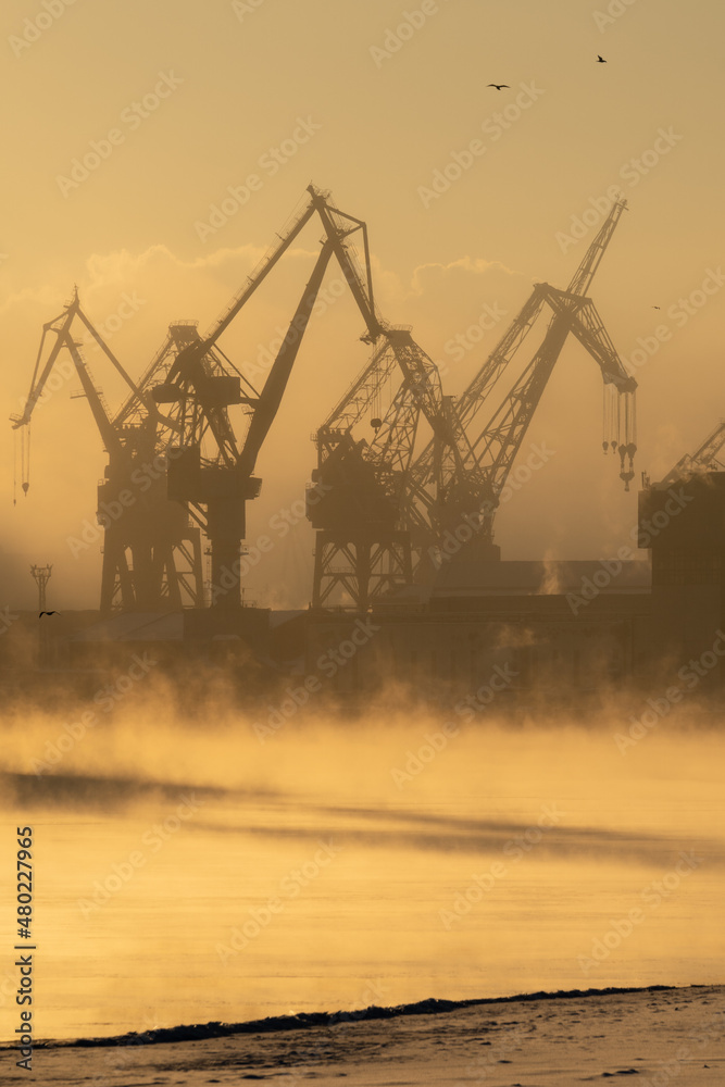 Cranes of Baltic shipyard in St. Petersburg in frosty winter day, steam over the Neva river, smooth surface of the river at sunset. 