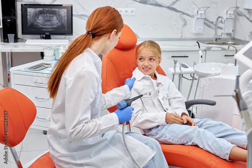 Dentist in gloves holding dental drill while treating child girl patient in modern dental office clinic  side view on adorbale excited child girl sitting on couch waiting for treatment.