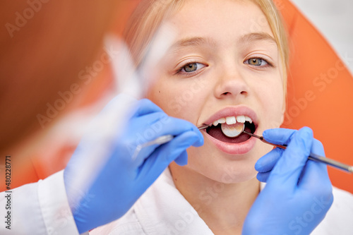 Close-up photo of child girl during dental tooth examination checkup. Attractive pretty caucasian kid sits with opened mouth while cropped female dentist in gloves examining inside of oral cavity
