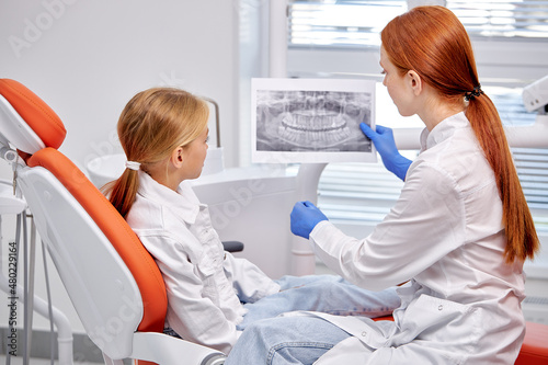 dental doctor showing oral x-ray exams oral radiography to child patient  in modern dental clinic. Side view on female dentist talking with kid girl  explaining how the treatment will be hold