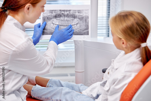 little Child girl sitting in dentist's chair looking at dental x-ray with doctor. Rear view on nice professional female Dentist showing x-ray to patient and explaining the procedure of treatment.
