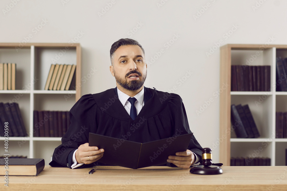 Judge passes judgement on the accused. Portrait of a young man in a robe reading the sentence while sitting at his table in the courtroom during a court session. Law, justice and legislation concept