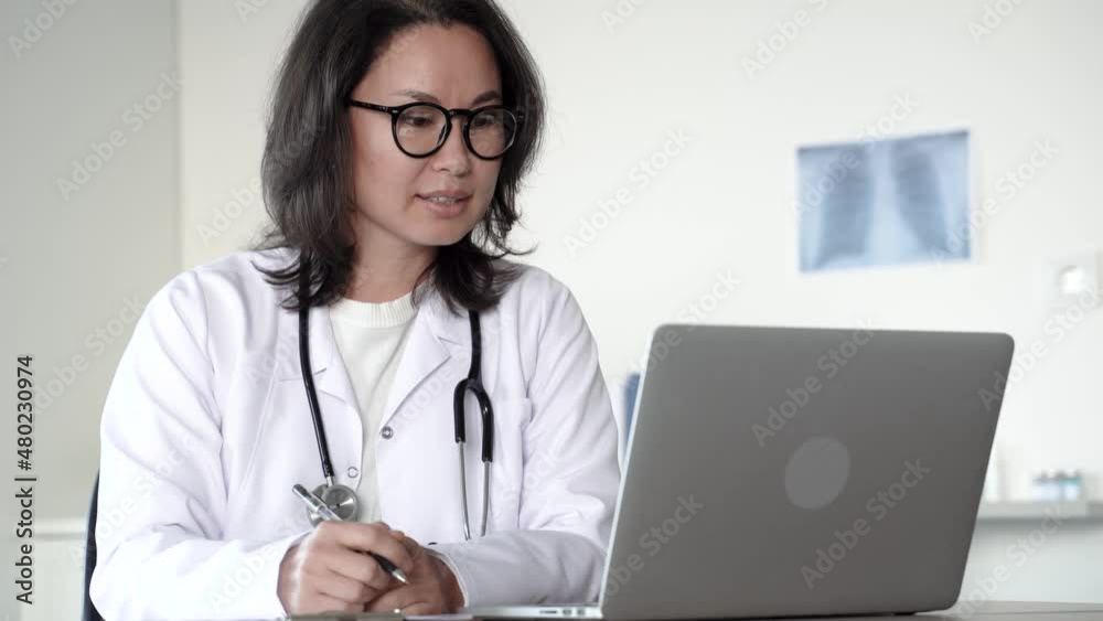 Attractive adult female asian doctor in lab coat, glasses and stethoscope using laptop at desk, looking at camera and talking with patient working in medical lab. distance consultation. 4k footage Attractive adult female asian doctor in lab coat, glasses and stethoscope using laptop at desk, looking at camera and talking with patient working in medical lab. distance consultation. 4k footage
