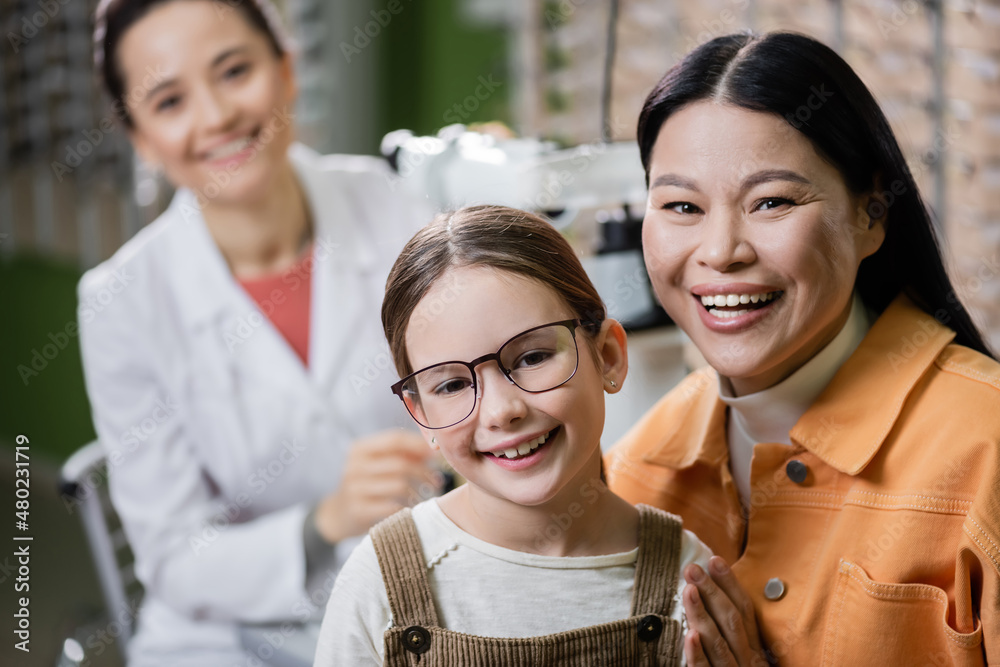 happy kid in eyeglasses looking at camera near asian mom and blurred ophthalmologist in optics shop. happy kid in eyeglasses looking at camera near asian mom and blurred ophthalmologist in optics shop.