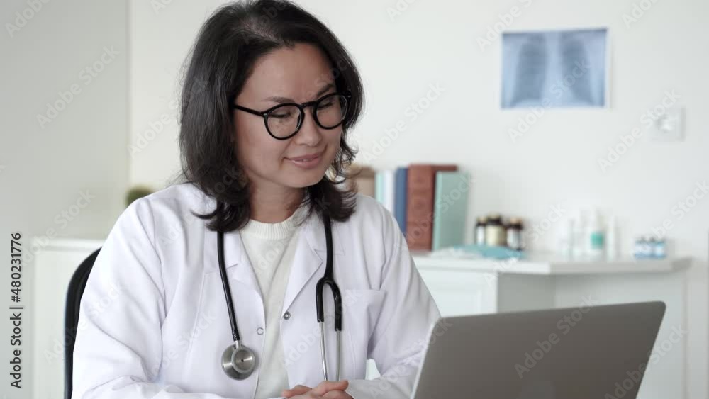 Attractive adult female asian doctor in lab coat, glasses and stethoscope using laptop at desk, looking at camera and smiling while working in medical office. distance consultation. 4k footage Attractive adult female asian doctor in lab coat, glasses and stethoscope using laptop at desk, looking at camera and smiling while working in medical office. distance consultation. 4k footage