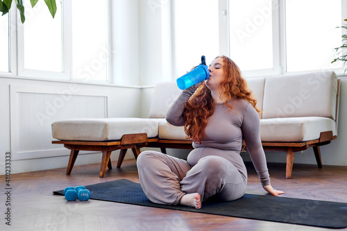 plus size woman with red hair siit on fitness mat drinking water, after training workout at home to lose weight and get strong flexible body, exercising on mat, photo