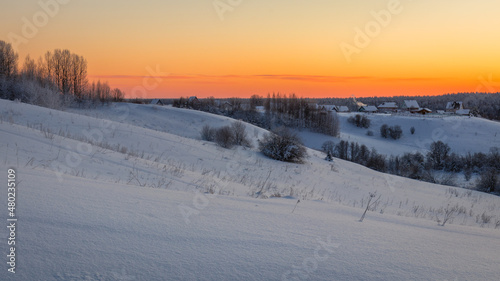 View of hills covered in snow with sparse trees and a small village against an orange sky. Travel concept © Aleksandr 44ARH