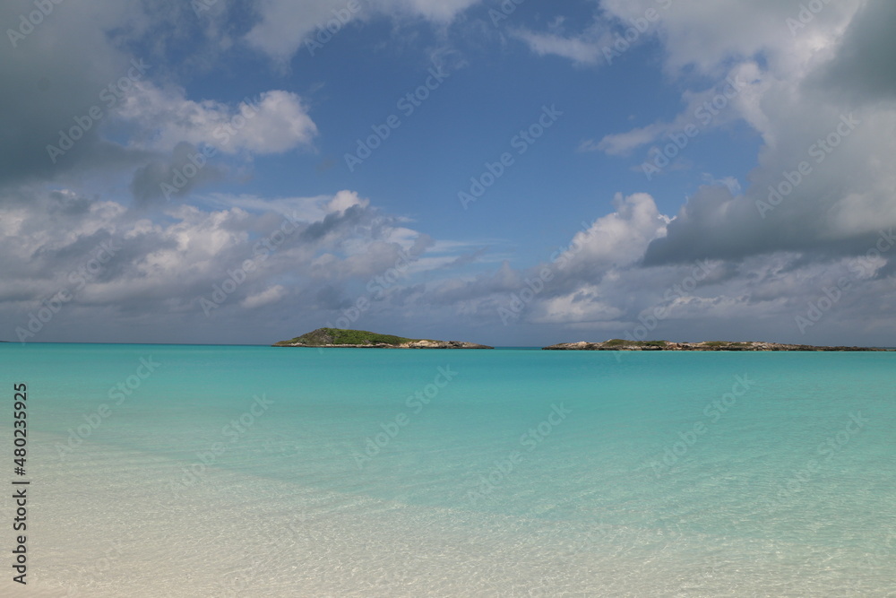 Fototapeta premium Exuma Bahamas white sand beach with blue skies, white clouds, small cays in the background. Blue green, turquoise, and relaxing water invokes calm during vacation. Deserted, secluded romantic beach.