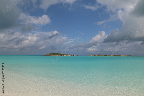 Exuma Bahamas white sand beach with blue skies, white clouds, small cays in the background. Blue green, turquoise, and relaxing water invokes calm during vacation. Deserted, secluded romantic beach.