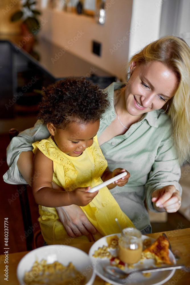 Cute child girl watching video cartoons on smartphone while having meal with mother, free time, leisure in the morning. pleasant caucasian female and black child enjoying time together