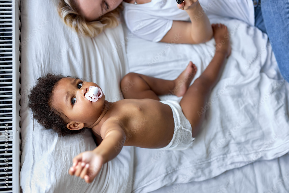 top view on cute child girl with black skin lying on bed with mother, with baby pacifier in mouth. view from above on shirtless kid after waking up in the morning. children, lifestyle concept
