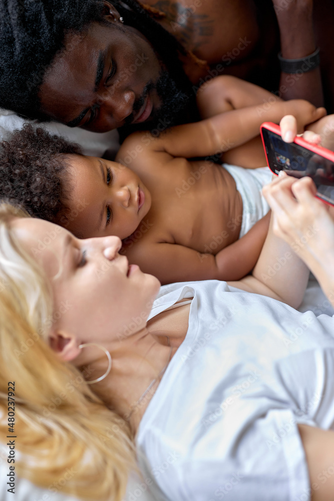 father mother and kid girl in diaper playing smartphone on bed in bedroom. Loving diverse Parents and little black child are resting at home. View from above. leisure, weekends. focus on child