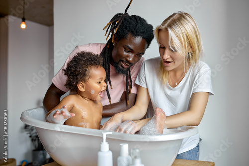 Konstfotografi Nice black man and blonde female washing daughter in bath, have fun, talking, explaining, playing with toys