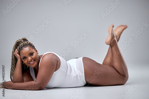 Positive Emotions Concept. Portrait of excited chubby black woman smiling, laughing looking at camera while lying on floor, wearing white bodysuit. Emotional black lady isolated on white background