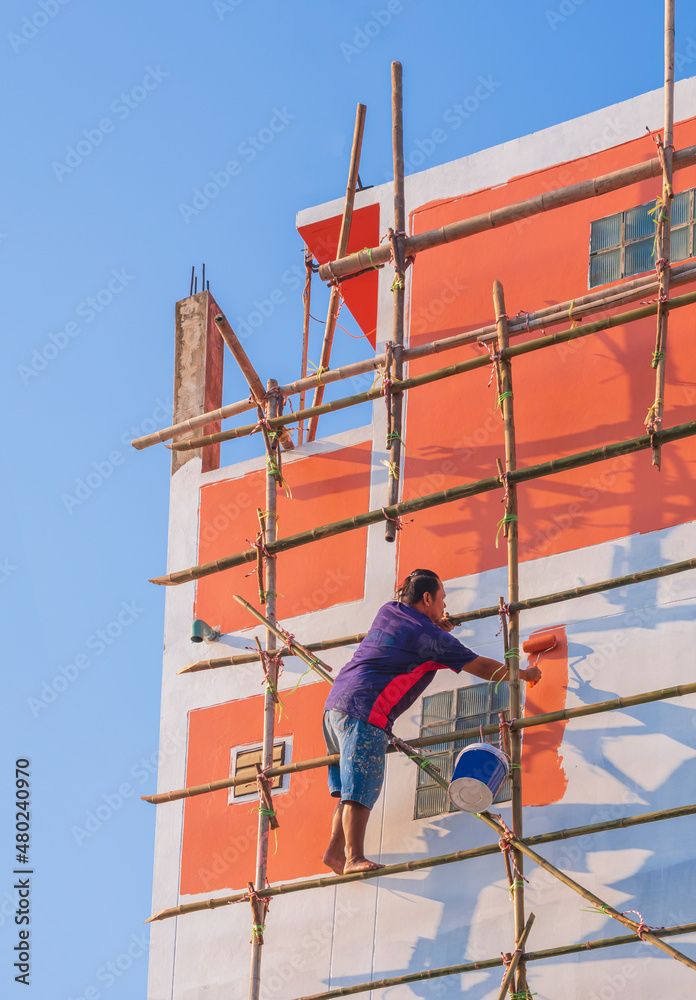 Asian builder worker on wooden scaffold is painting the old wall outside of house against blue clear sky in vertical frame