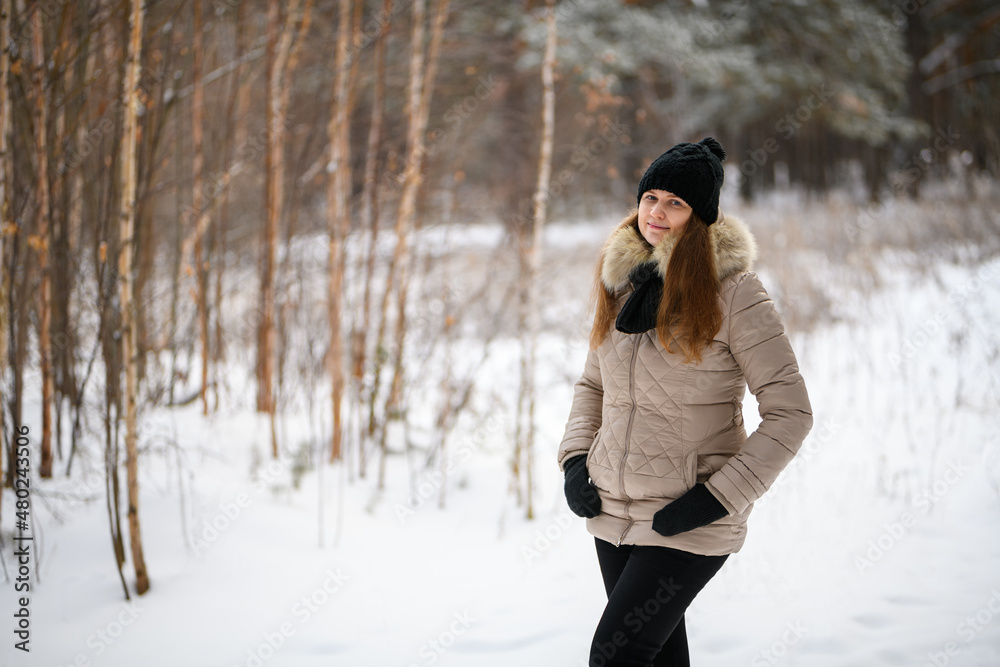 Portrait of a red-haired girl in winter forest in warm clothes: black hat and light jacket looking at the camera. Blurred forest background Portrait of a red-haired girl in winter forest in warm clothes: black hat and light jacket looking at the camera. Blurred forest background