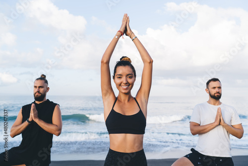 Portrait of cheerful female yogi in sportswear smiling at camera during together holistic retreat at seashore beach  happy woman with slim figure posing during yoga practice with men at coastline