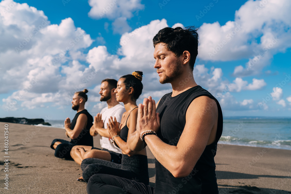 Young male and female in lotus pose holding hands in namaste enjoying spiritual meditation at coastline beach, group of friends have togetherness morning yoga practice at seashore in Indonesia Young male and female in lotus pose holding hands in namaste enjoying spiritual meditation at coastline beach, group of friends have togetherness morning yoga practice at seashore in Indonesia