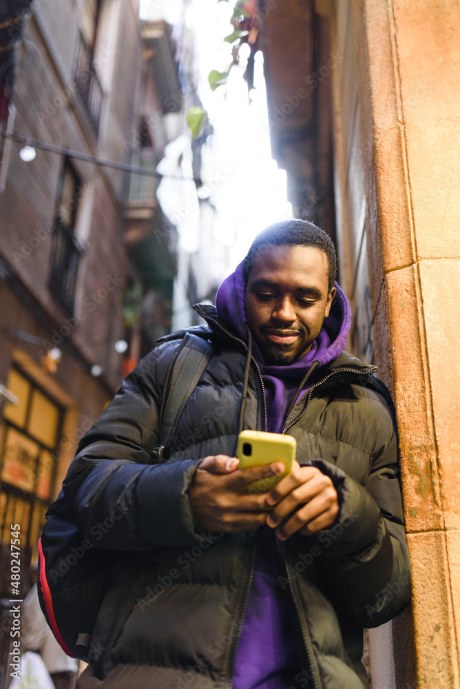 Low angle of African American man in casual outerwear with backpack. He is leaning on building wall and browsing social media on mobile phone on city street