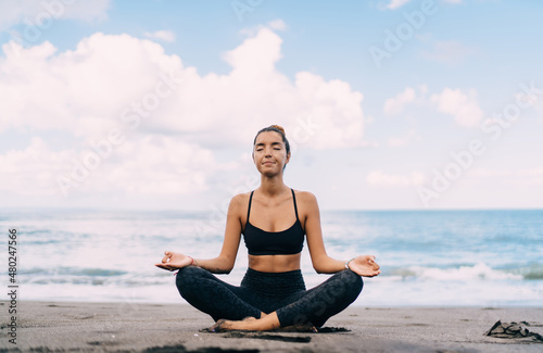 Middle Eastern fitness girl sitting asana for training vitality wellness and tranquility, slim woman in casual sportive wear recreating on meditation in lotus pose practice yoga for health at seashore