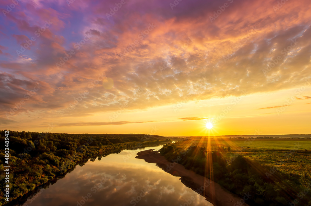 Scenic view at beautiful sunset or sunrise on a shiny river with green bushes on sides, golden sun rays, calm water ,deep blue cloudy sky and forest on a background, spring landscape