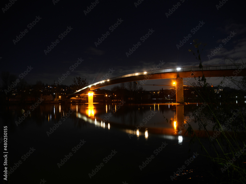 Beleuchtete gebogene Fußgängerbrücke bei Nacht über einen Fluss mit Bäumen am Ufer